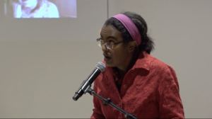 Dark skinned woman in coral shirt, pink headband, and glasses speaking at a podium
